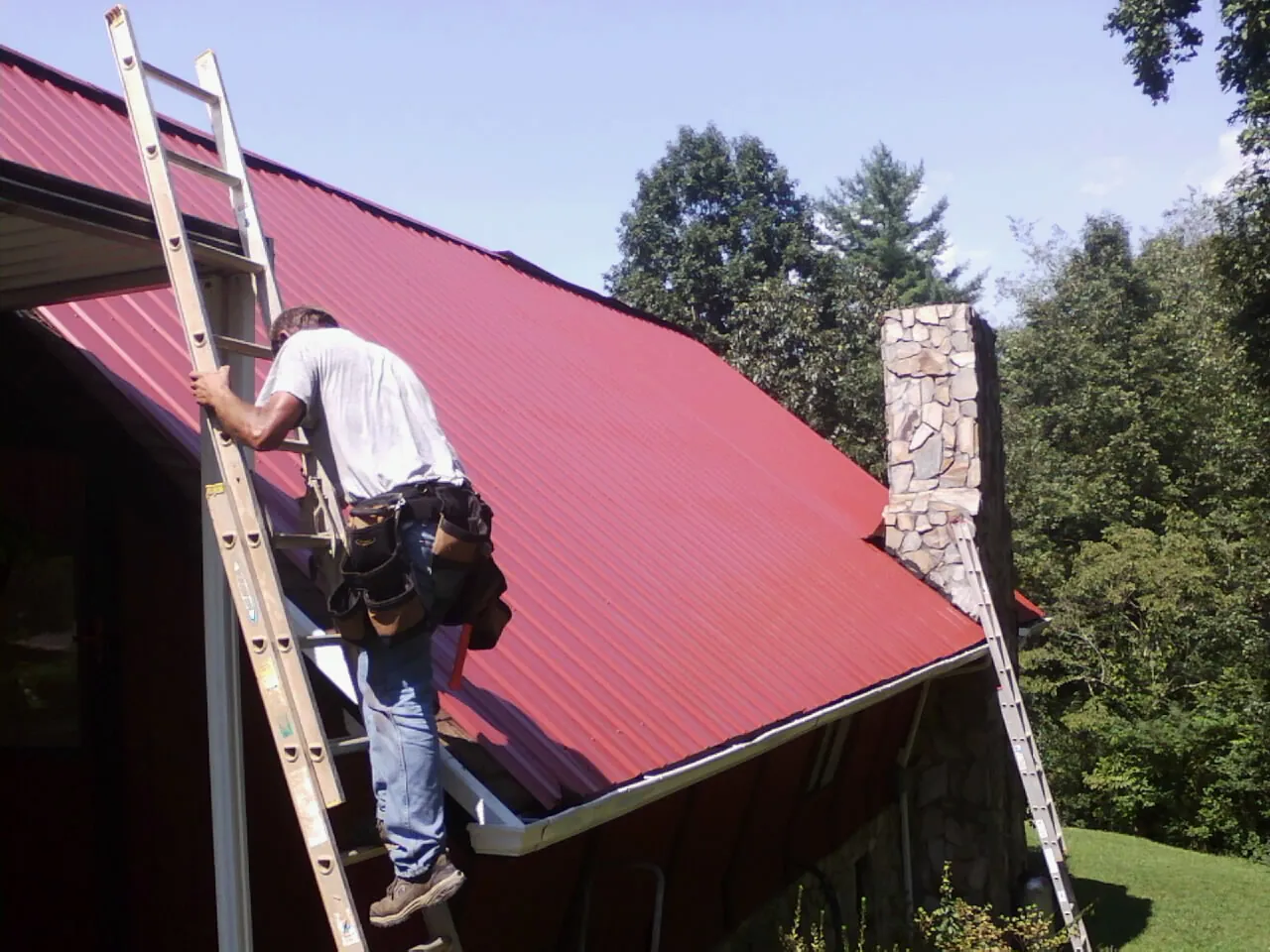 Roofer on a ladder beside a red metal roof and stone chimney — Soffit & Fascia Repair by Roofing Brothers in Mooresville